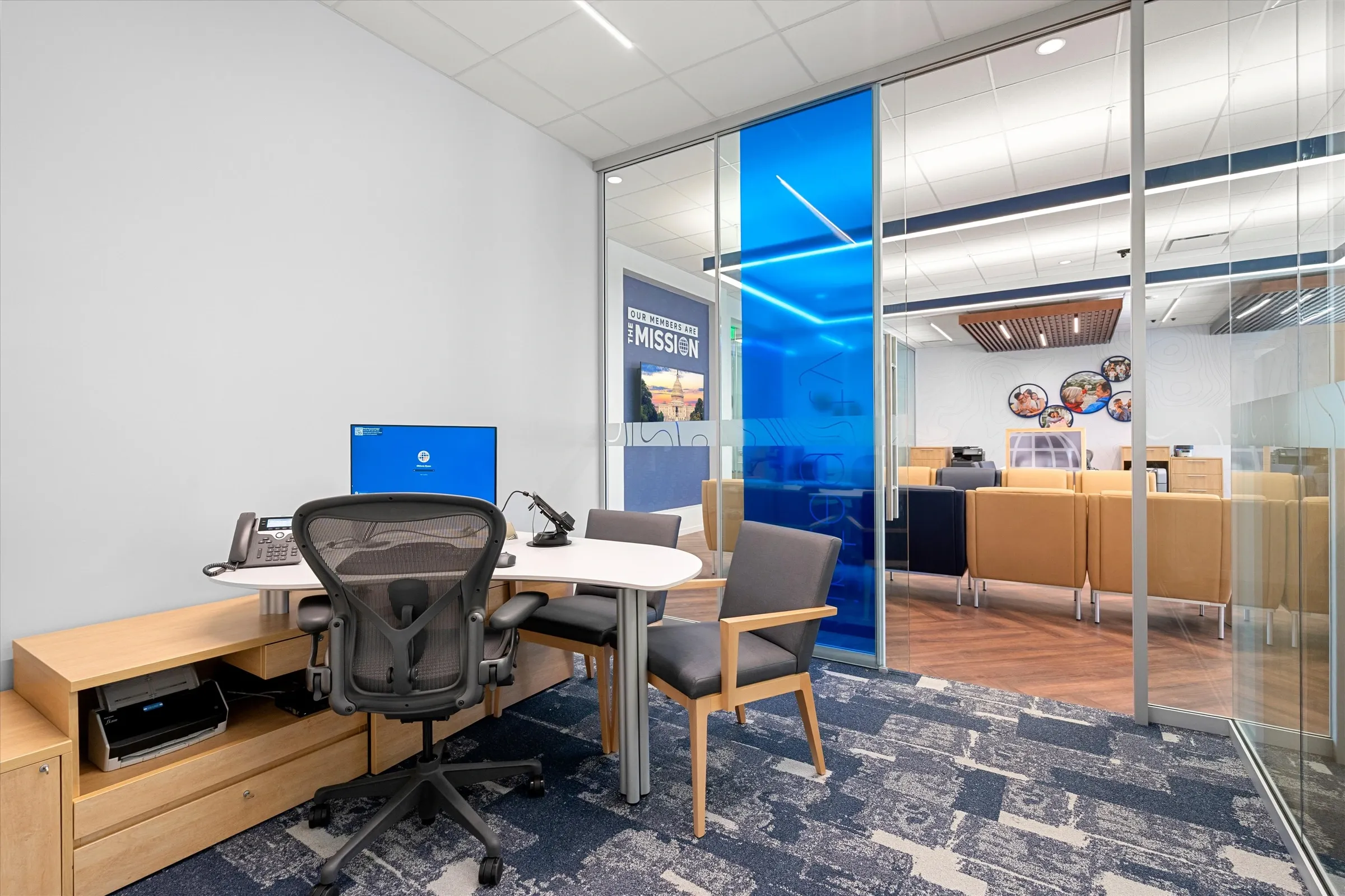 Glass-walled consultation office with ergonomic desk, guest chairs, blue branded "Mission" panel visible through glass, and blue patterned carpet