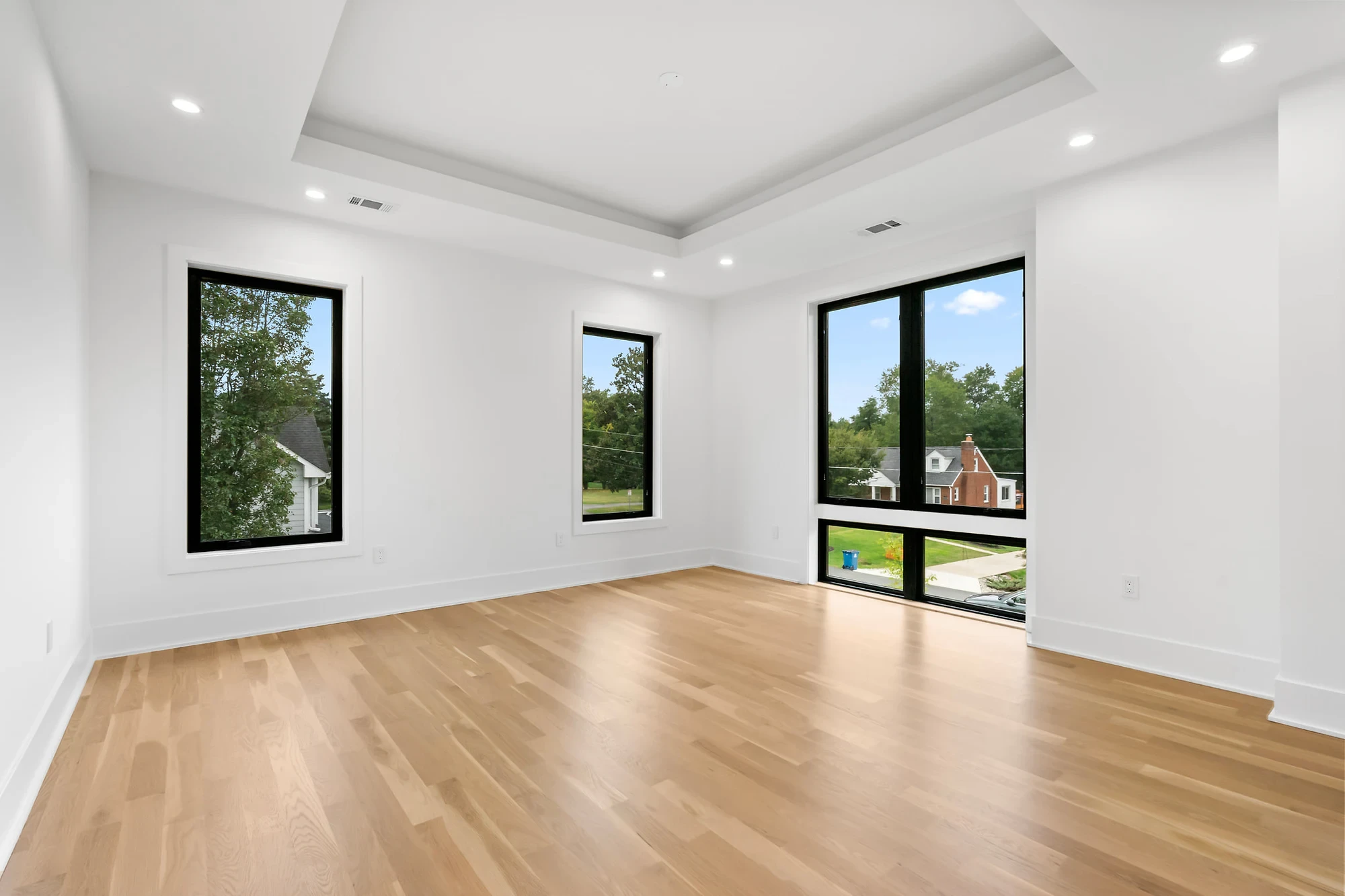 Vacant new-construction living room with a tray ceiling, white oak floors, and black-framed corner windows — before virtual staging