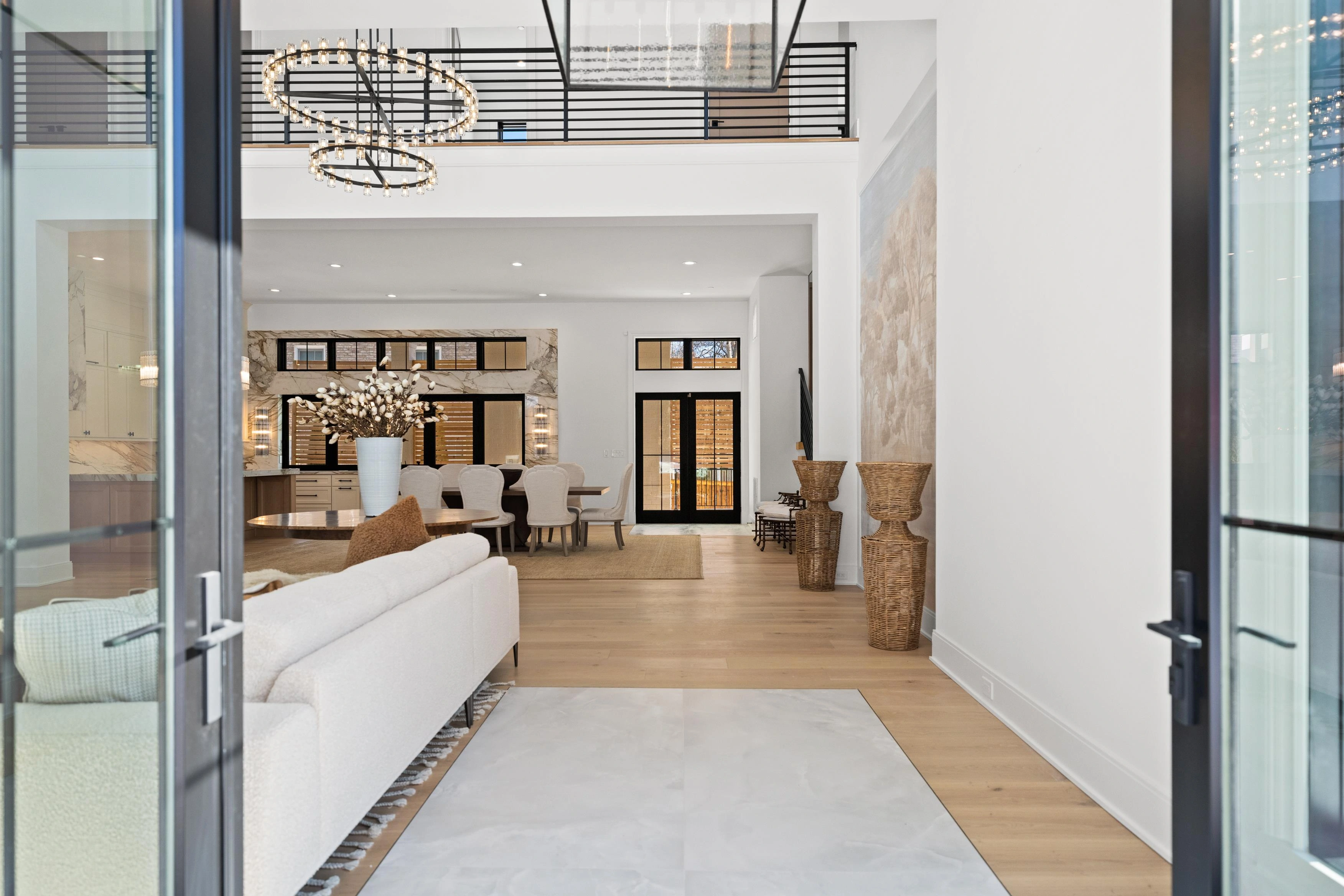 Double-height foyer with ring chandelier, open to living and dining areas through glass entry doors
