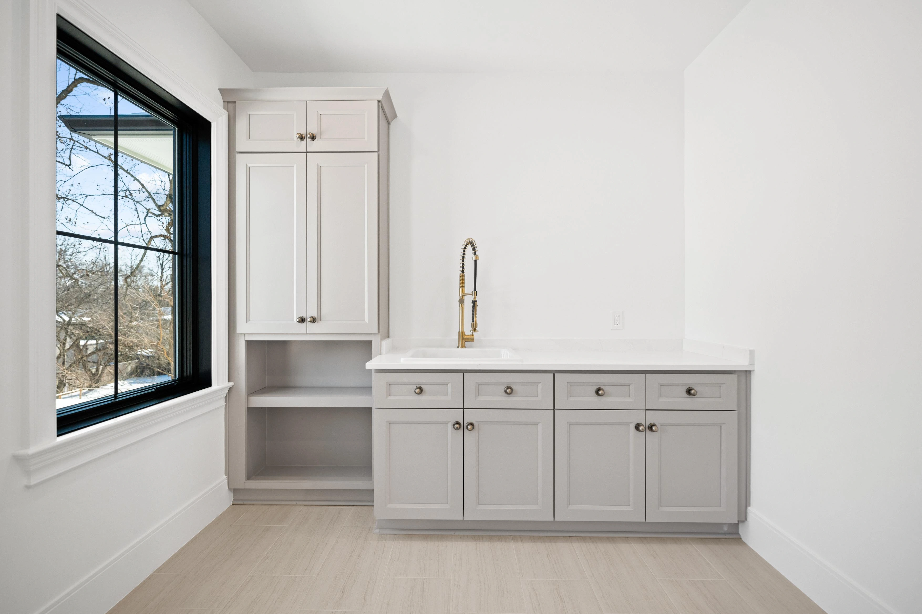 Laundry room with grey shaker cabinetry, gold commercial-style faucet, and black-framed window