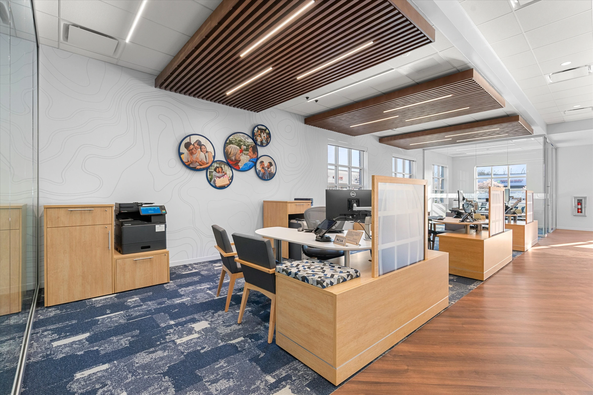 Advisor workstation area with oak privacy-panel desks, circular military family wall art, wood slat ceiling accents, blue patterned carpet, and printer station