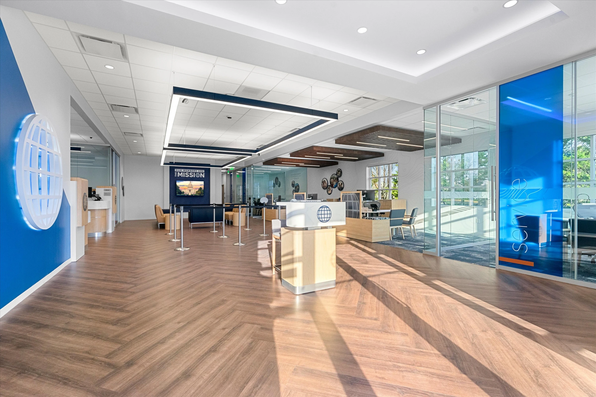 Wide branch floor view with "Our Members Are The Mission" display, stanchion queue, advisor desks, wood slat ceiling accents, and glass consultation rooms