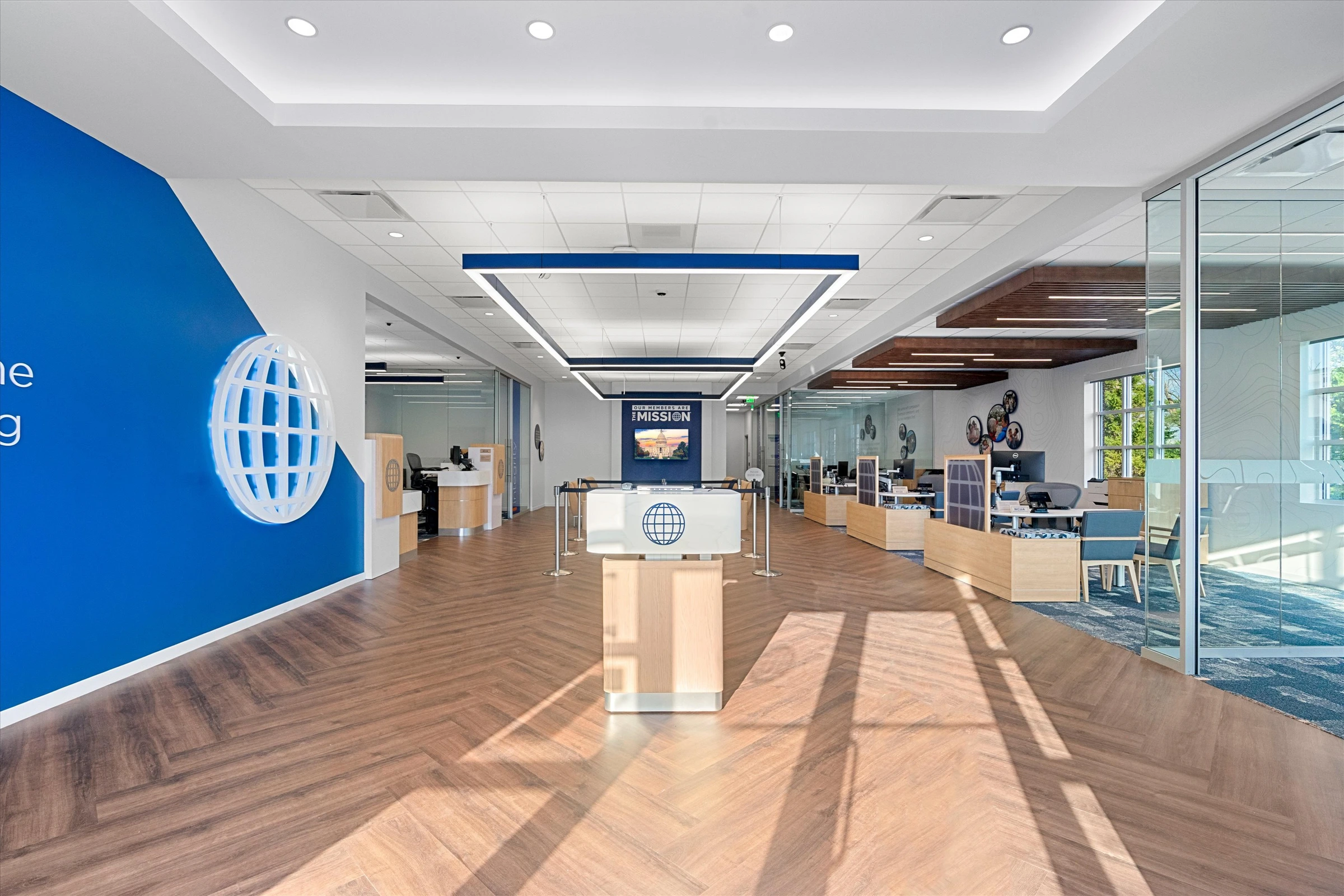 Full branch interior perspective from entrance showing herringbone flooring, greeter podium, advisor workstations, glass offices, and natural light
