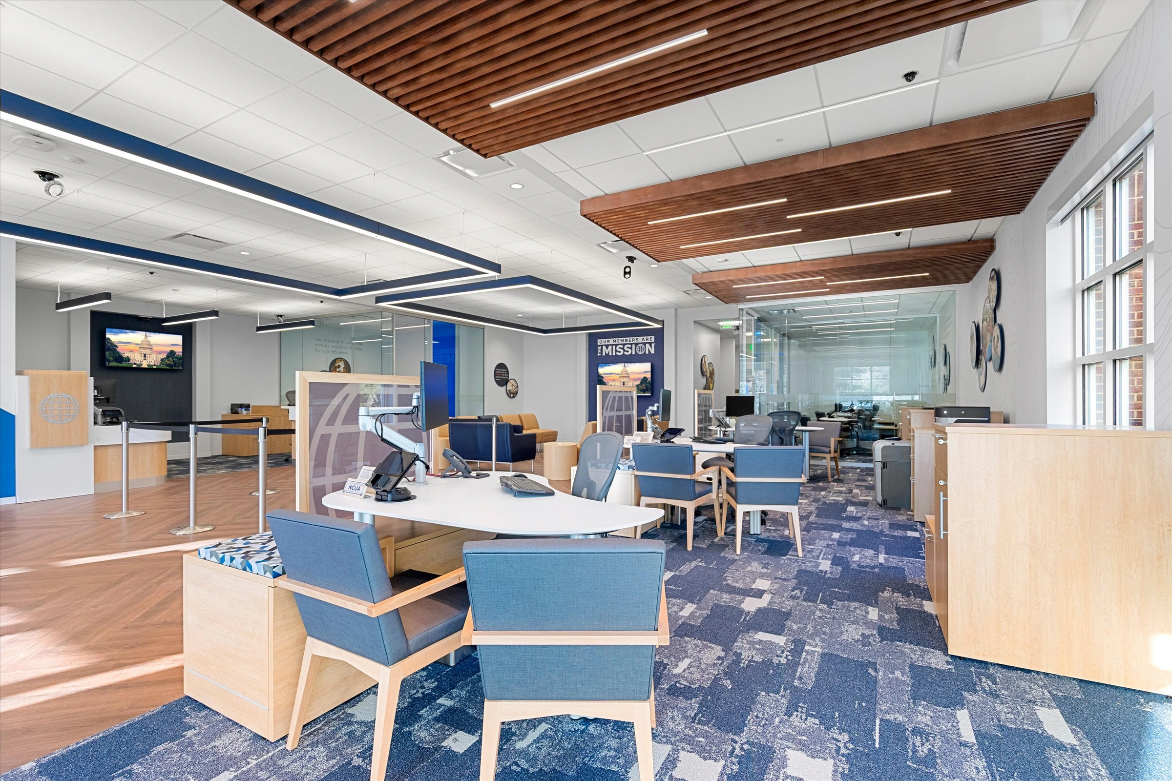 Open branch floor from advisor area showing greeter desk, blue branded wall, stanchion queue, member seating, and wood slat ceiling details