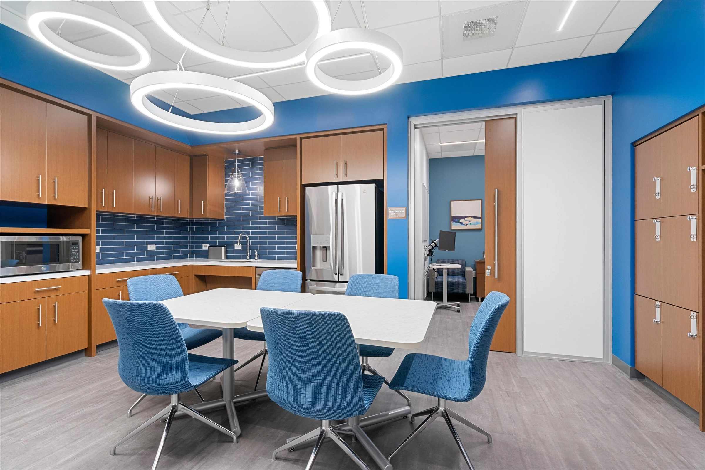Employee break room with blue accent walls, modern ring LED ceiling lights, blue subway tile backsplash, oak cabinetry, stainless appliances, and blue upholstered dining chairs