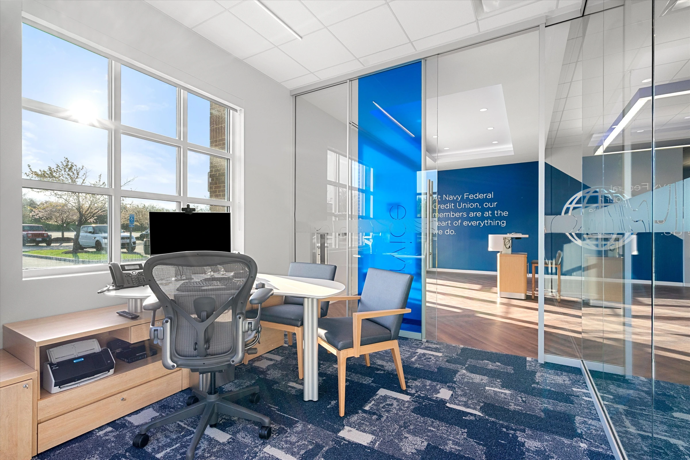 Corner consultation office with large windows, natural sunlight, ergonomic desk setup, guest chairs, blue branded glass partition, and parking lot view