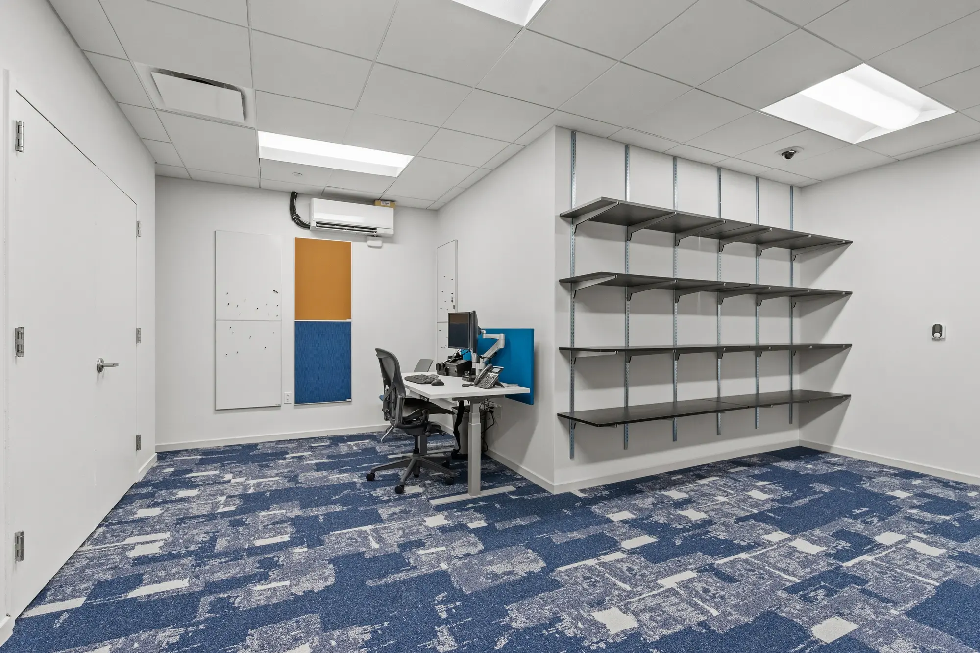 Back-office workstation room with blue patterned carpet, dual monitors, metal document shelving, and colorful pinboard