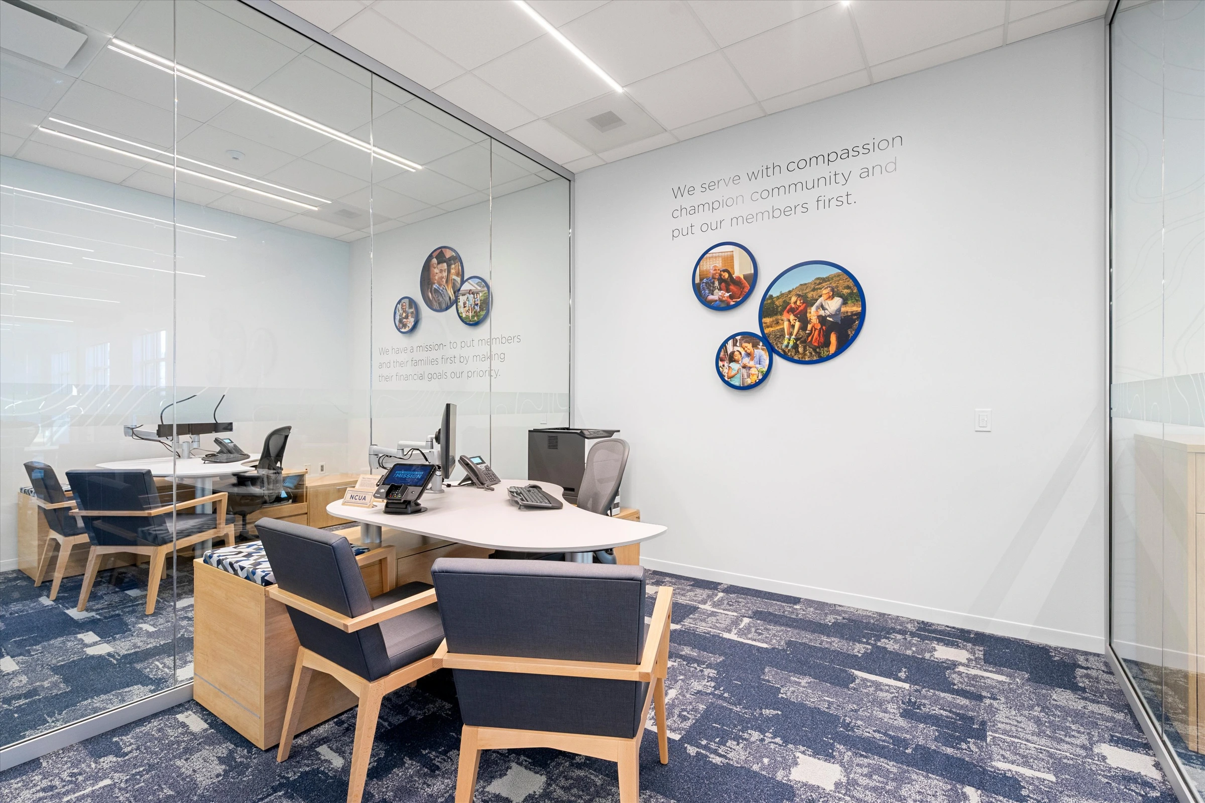 Private member office with "We serve with compassion" wall text, circular military family photo displays, glass partition, desk with monitor, and blue carpet