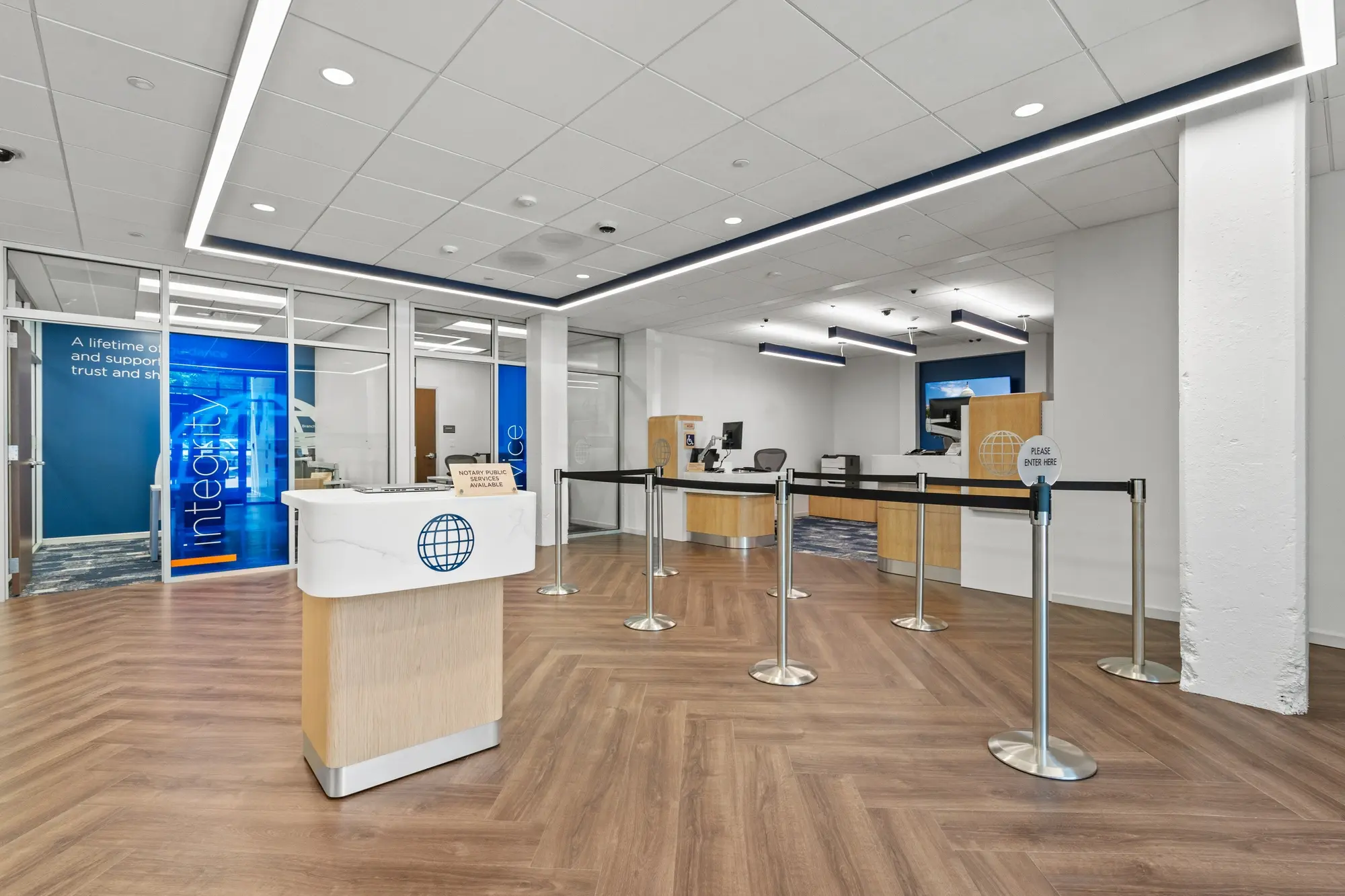 Reception area with branded greeter podium, stanchion queue, glass integrity room, and teller counter in background