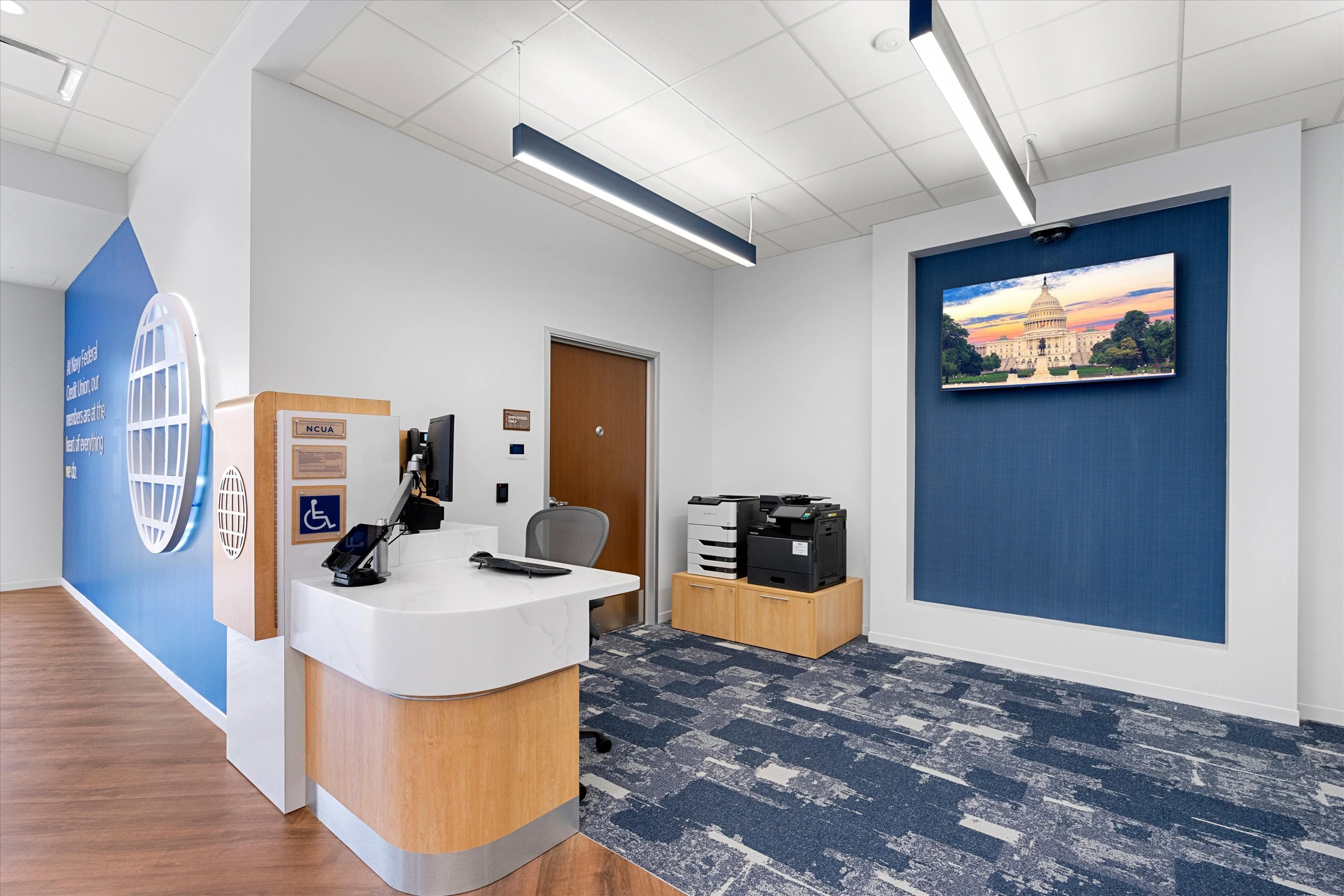 Reception area with branded globe-logo greeter desk, navy accent wall with Capitol display, accessibility signage, and printer station