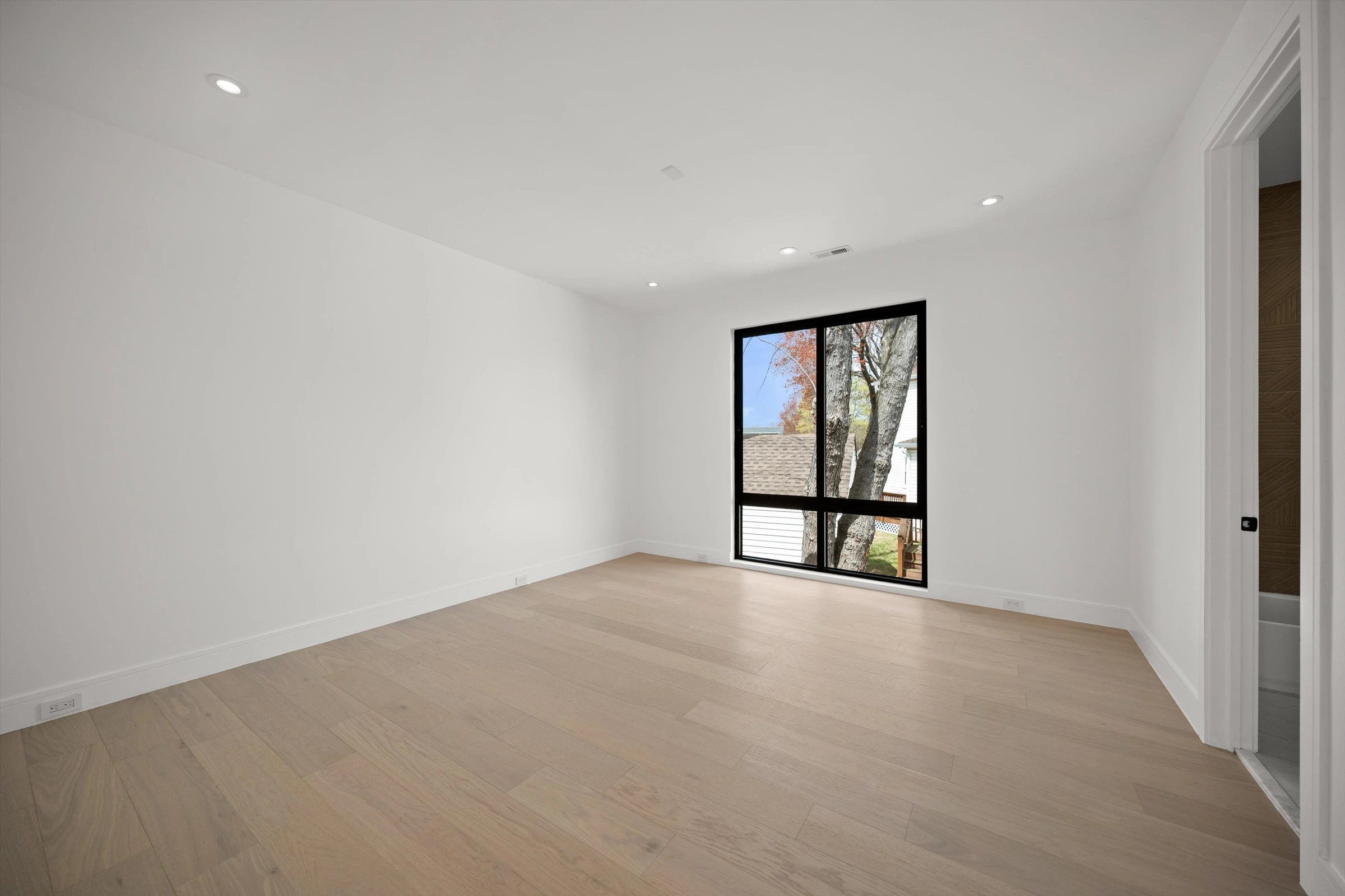 Second bedroom with large black-framed window, light oak hardwood floors, recessed lighting, and tree views