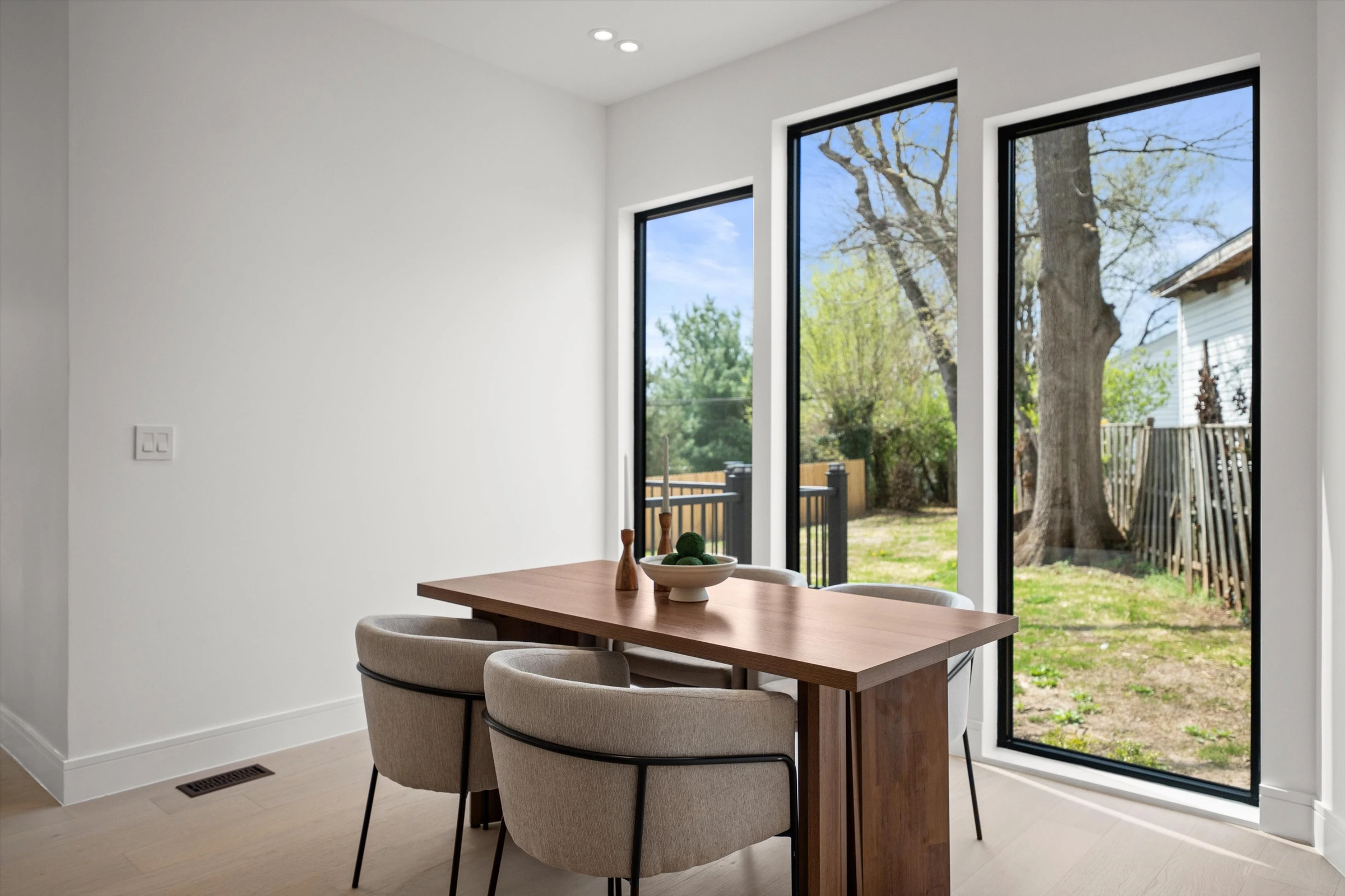 Casual dining area with walnut table, curved gray chairs, and floor-to-ceiling windows overlooking backyard deck and mature trees