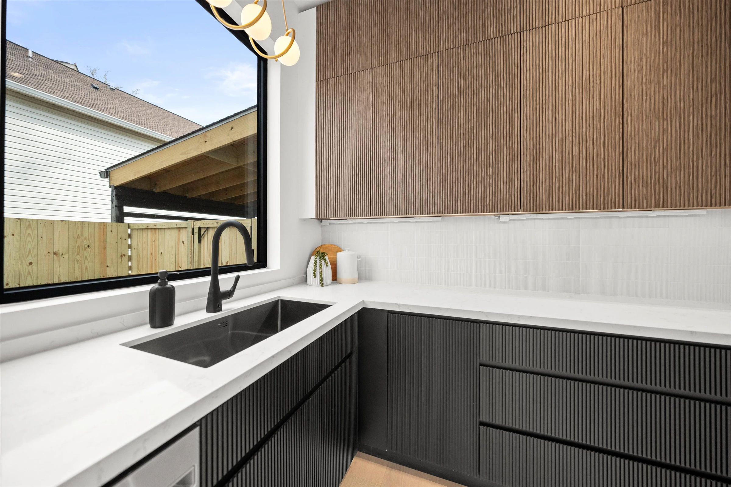 Kitchen butler sink detail with matte black faucet, white quartz countertop, walnut reeded upper cabinets, and window overlooking carport