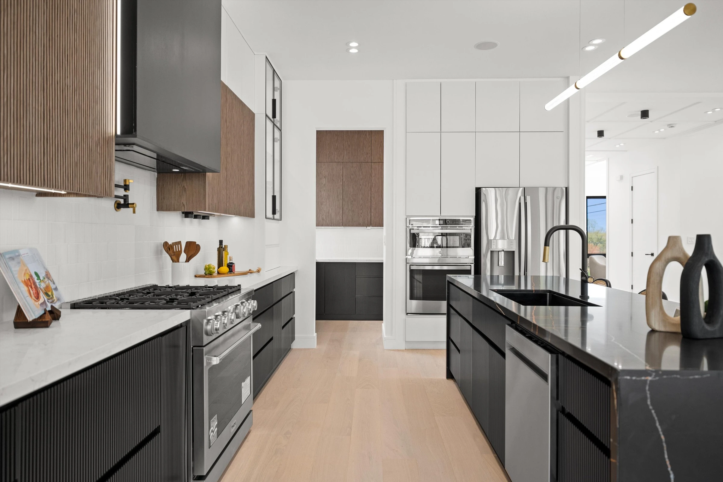 Kitchen galley view with professional gas range, gray range hood with walnut surround, dark fluted lower cabinets, white quartz countertops, and stainless appliances