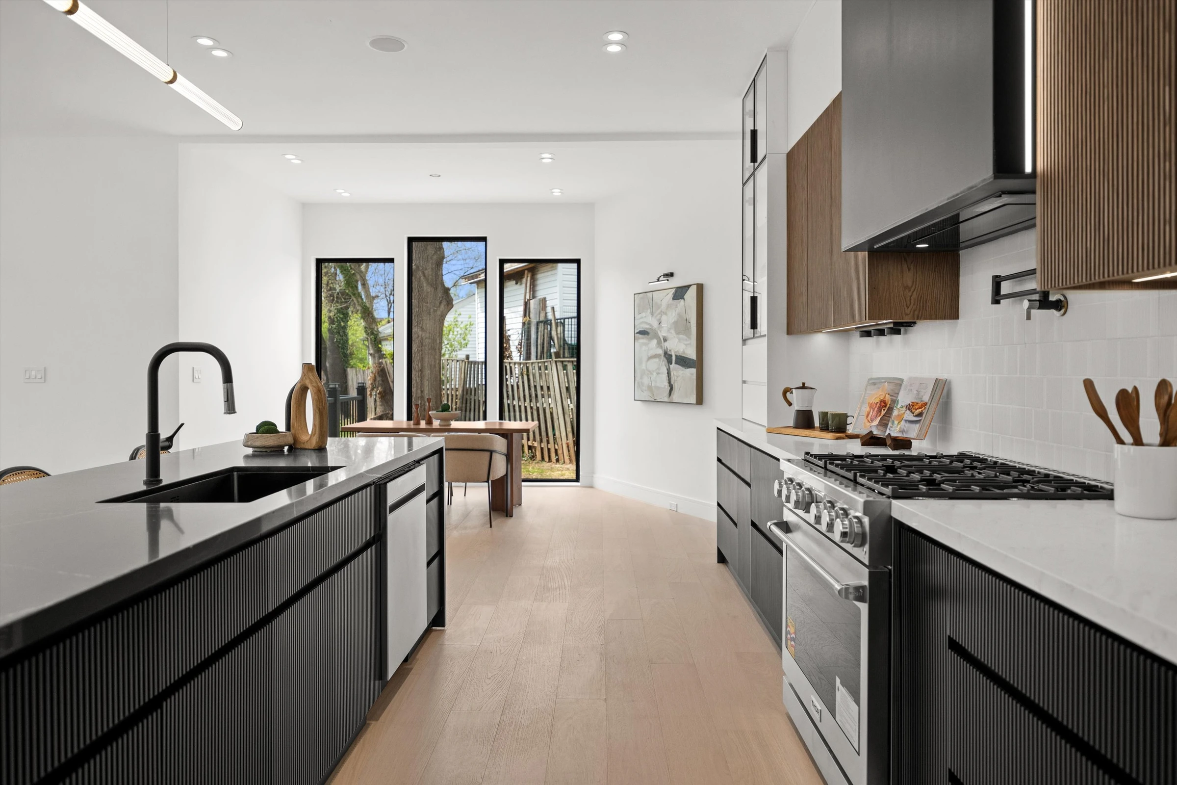 Kitchen toward dining nook with black island, gas range, walnut hood surround, and floor-to-ceiling windows looking out to backyard