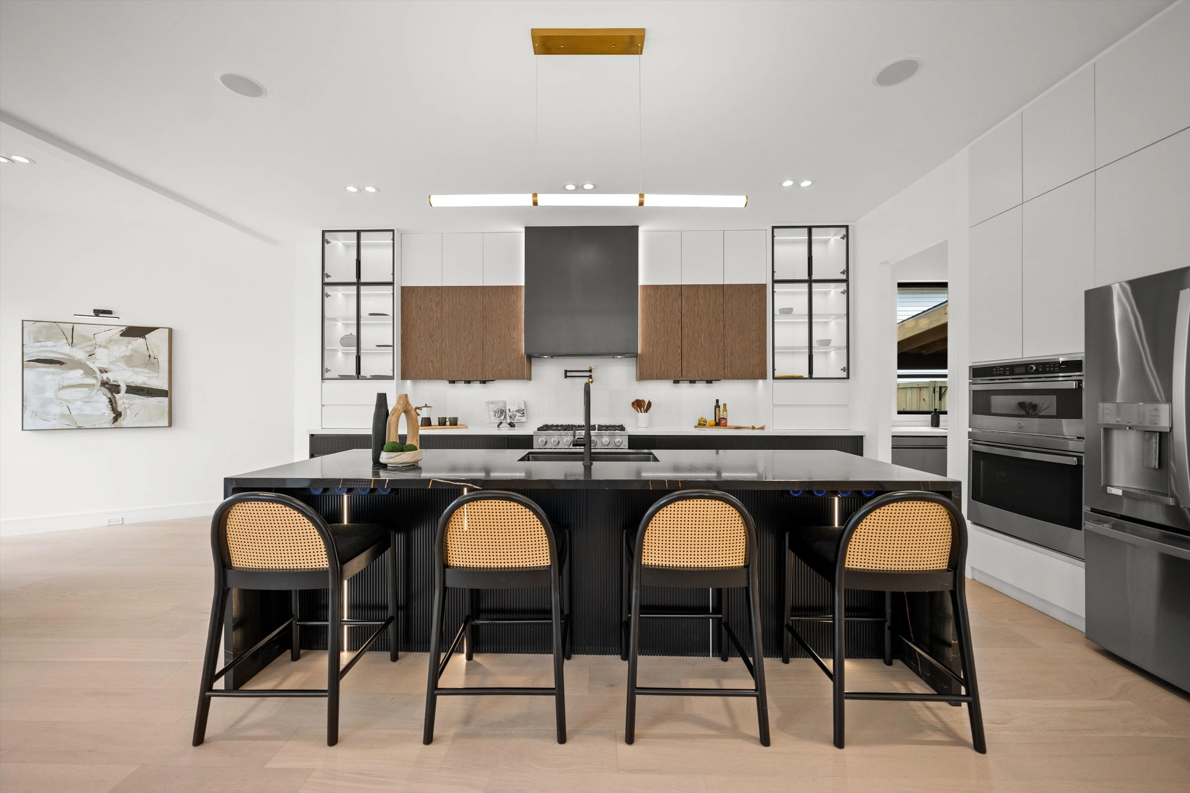 Designer kitchen with black marble waterfall island, cane-back bar stools, two-tone cabinetry, wood-accented range hood, and gold pendant light