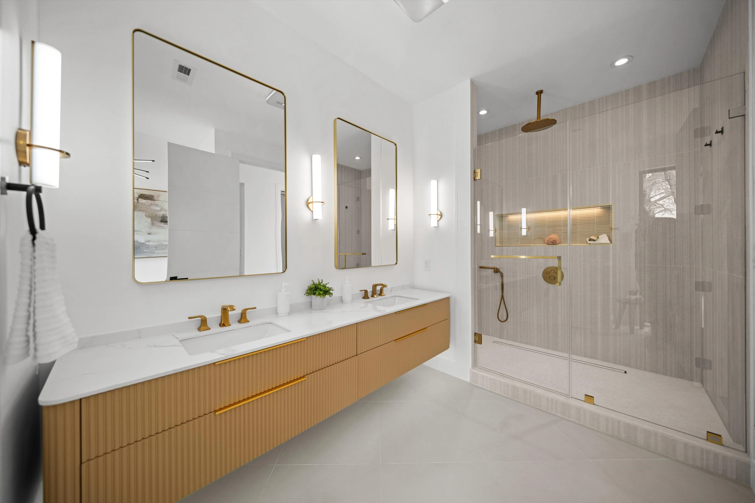Primary bathroom with floating gold fluted vanity, dual sinks, gold fixtures, frameless glass rain shower with linear tile, and illuminated mirrors
