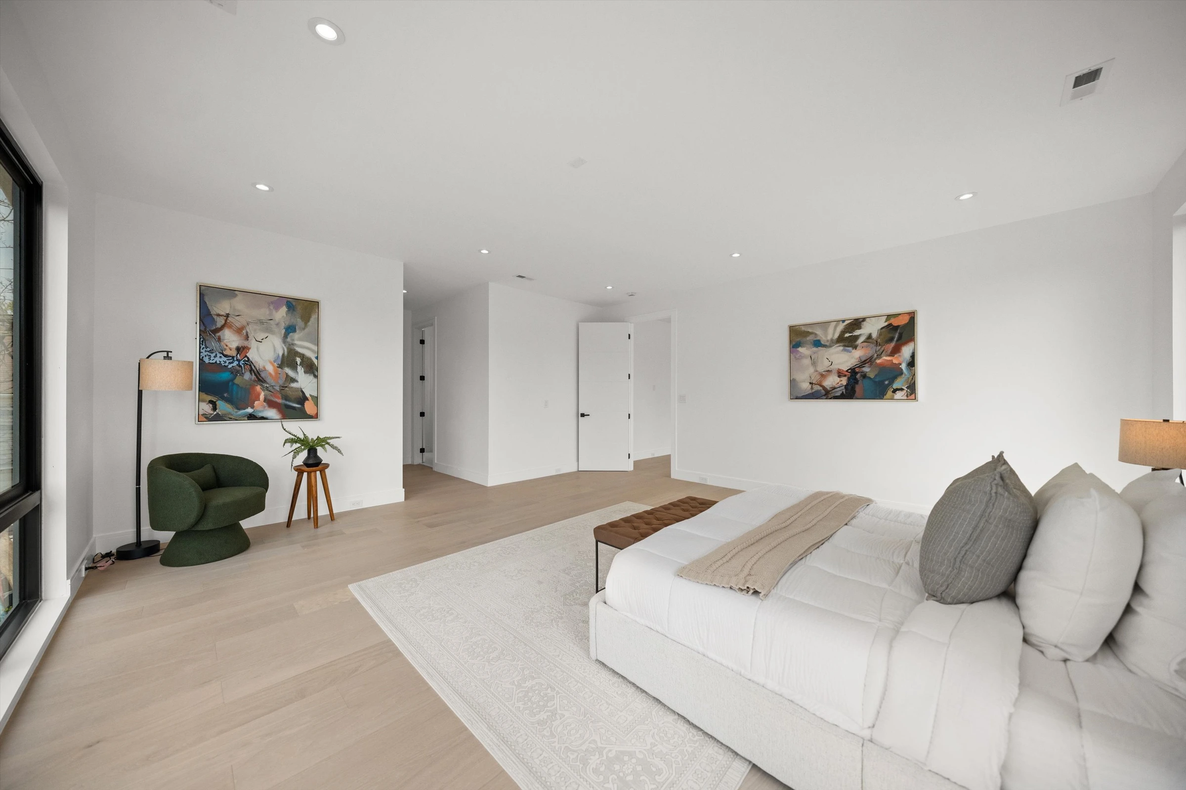 Primary bedroom alternate angle showing colorful abstract artwork, green accent chair, generous floor space, and natural light