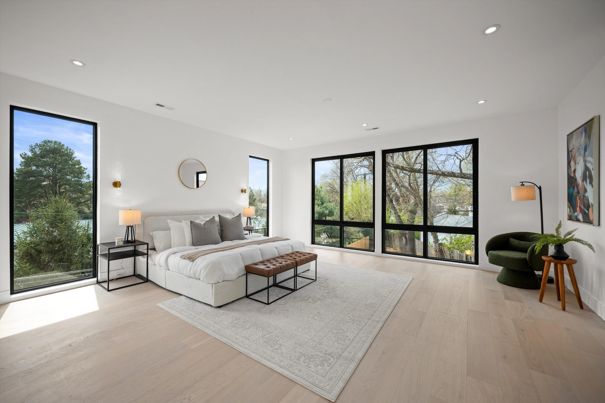 Spacious primary bedroom with floor-to-ceiling black-framed windows on two walls, light oak floors, upholstered bed, leather bench, and treetop views