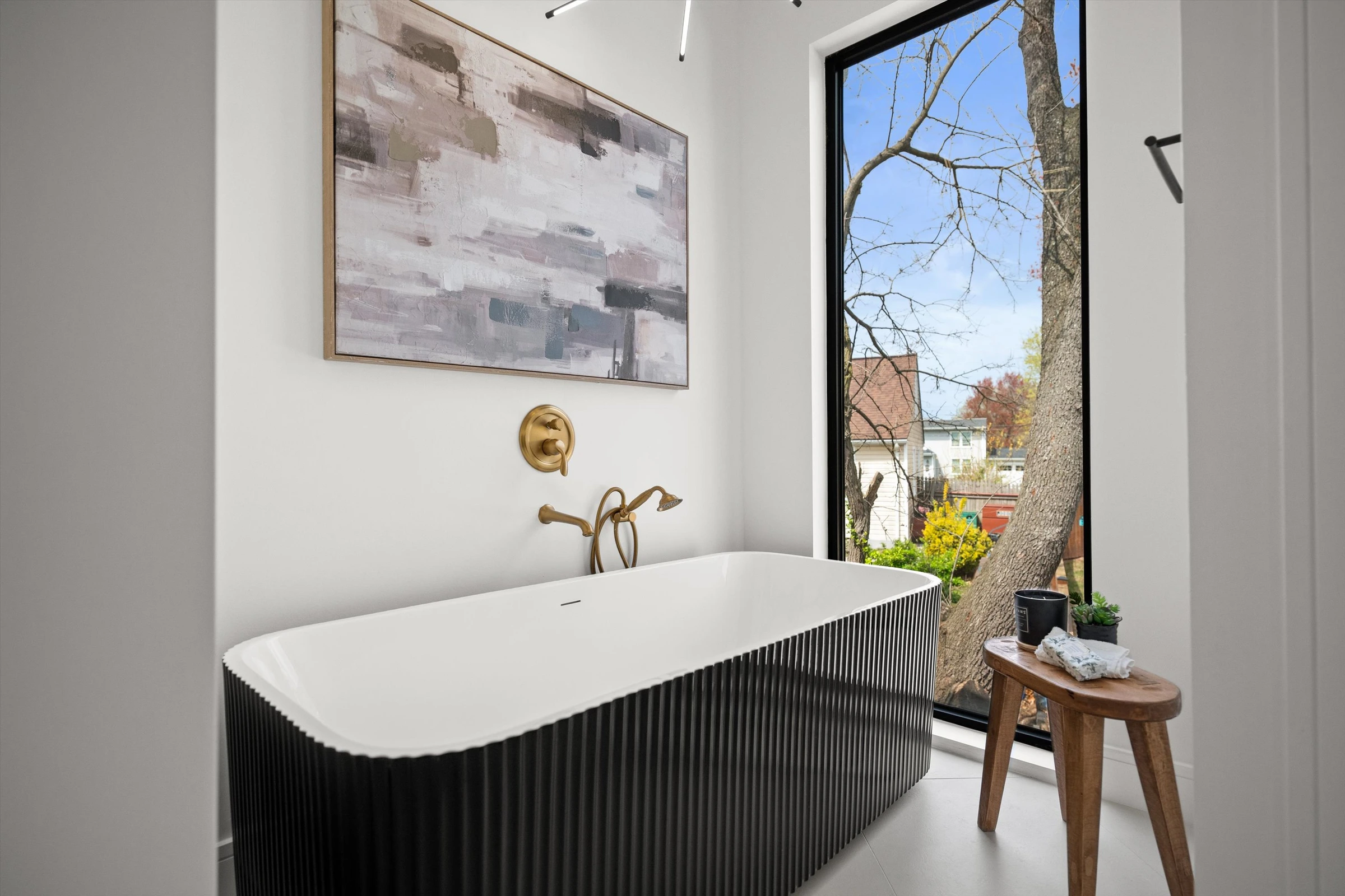 Freestanding black fluted soaking tub with gold wall-mount faucet, abstract artwork, and floor-to-ceiling window with garden view