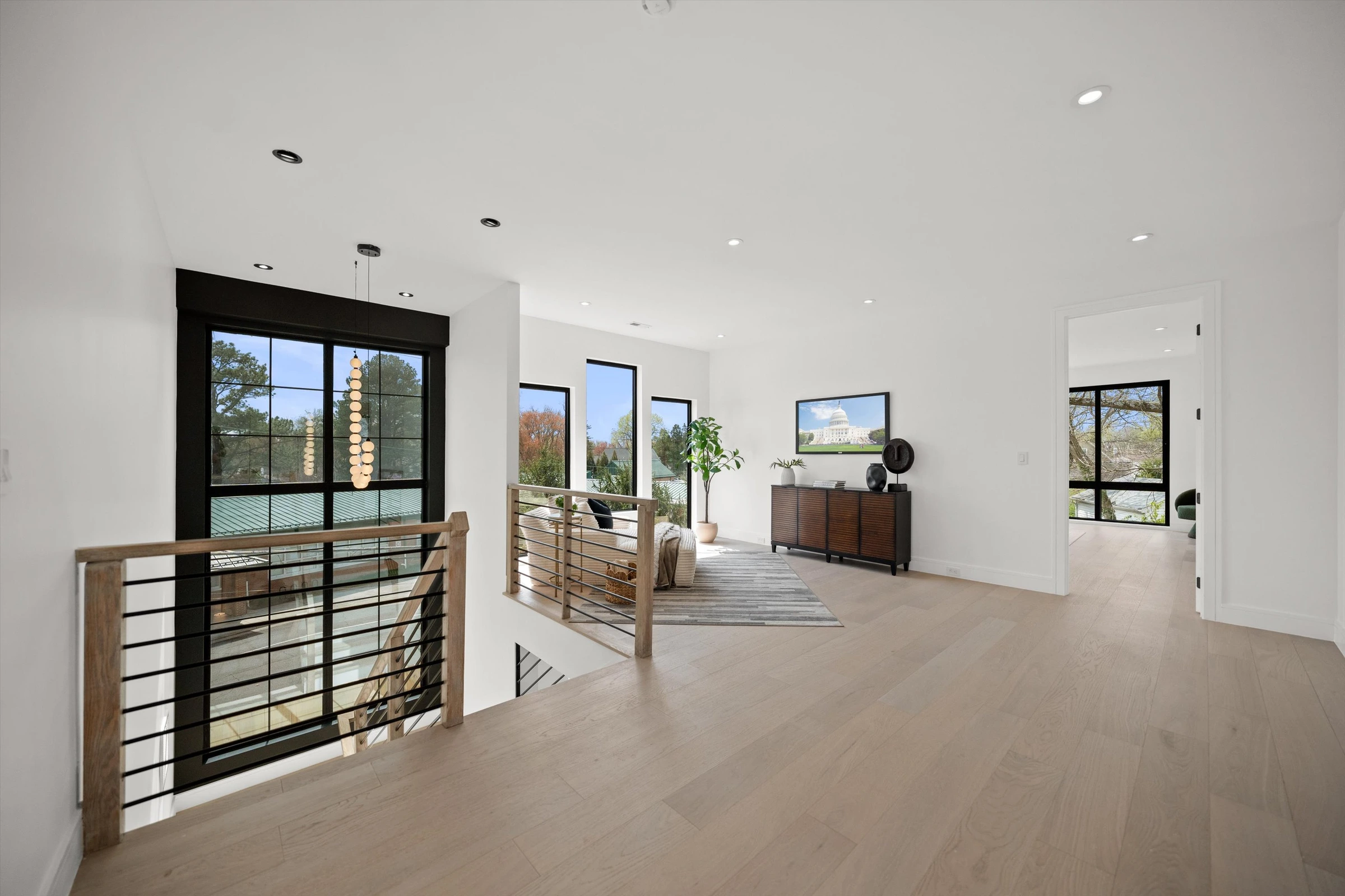 Second-floor hallway with double-height window overlooking entry, open railing, light oak floors, and modern credenza with art