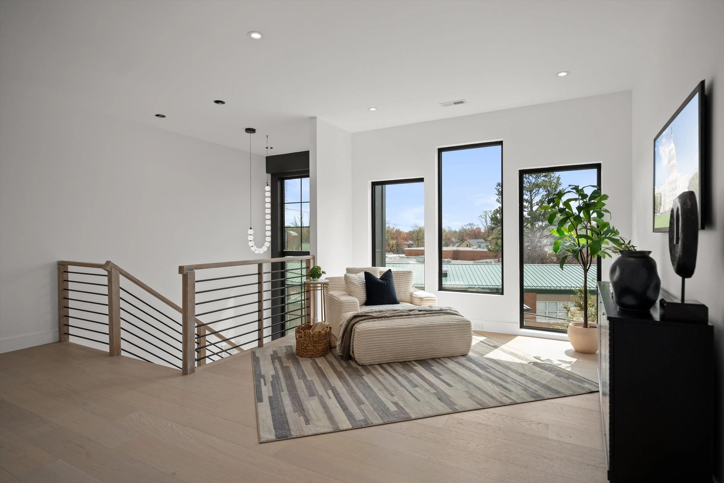 Upper-level landing with horizontal metal railing, lounge chaise, tall windows with neighborhood views, and pendant light over stairwell